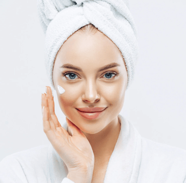 A smiling woman in a white bathrobe applying facial moisturizer cream for a glowing skincare routine.