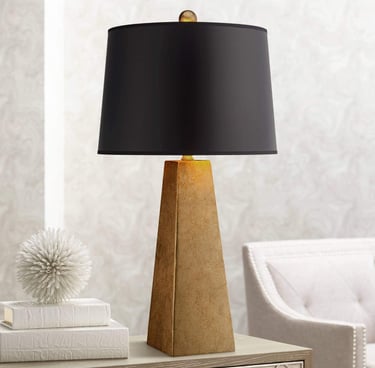 Modern gold pyramid table lamp with a black drum shade on a nightstand for bedroom lighting.