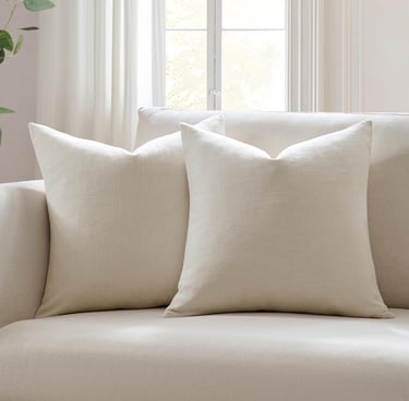 Two cream textured square throw pillows on a matching beige sofa in a bright living room.