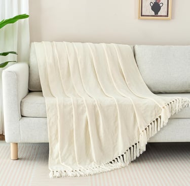Cream-colored woven throw blanket with fringe draped over a modern light gray sofa in a living room.