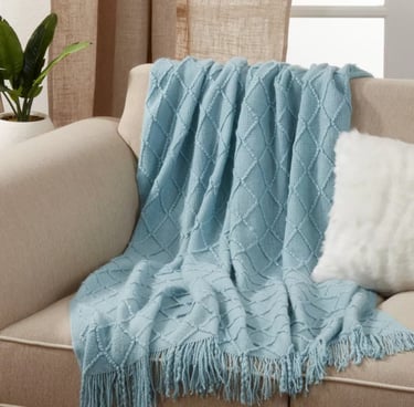 Light blue diamond pattern knitted throw blanket with fringe draped on a beige sofa.