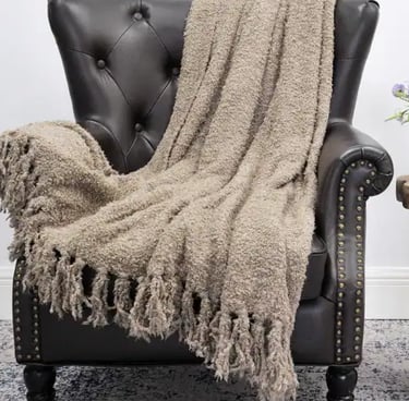 Cozy taupe textured throw blanket with tassels draped over a dark leather tufted armchair.