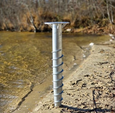 A galvanized steel helical ground screw anchor installed in the sandy shoreline of a lake for dock construction.