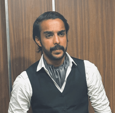 A man with a mustache wearing a black vest, cravat, and white shirt standing against a wood wall.