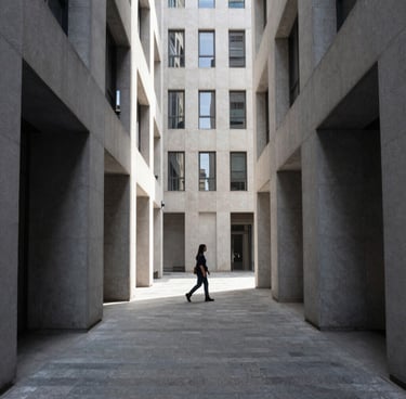 Candid street life photography in a clean, modern Western city, featuring a person walking through a shaft of light between tall buildings, high contrast, slate and light grey palette.