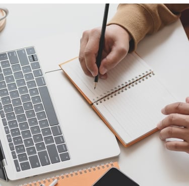 someone writing with a pen in a note book a laptop computer is on the desk