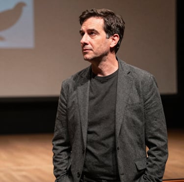 A professional portrait of a theater director in a North American / US setting, wearing refined charcoal attire, looking thoughtfully toward a stage with soft background lighting.