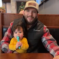 A smiling man in an Ariat vest and trucker hat holds a baby at a restaurant table.