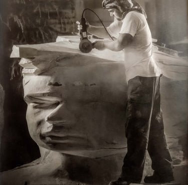 Artist using a power tool to carve a large stone head sculpture in a workshop.