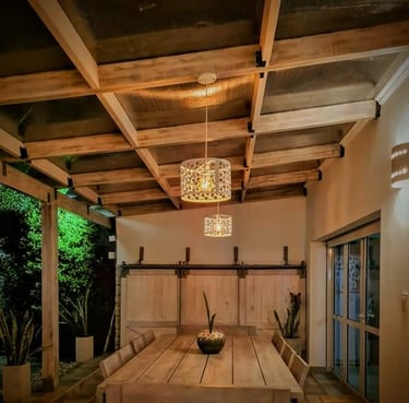 Comedor rústico al aire libre con una mesa de madera bajo una pérgola con iluminación colgante.