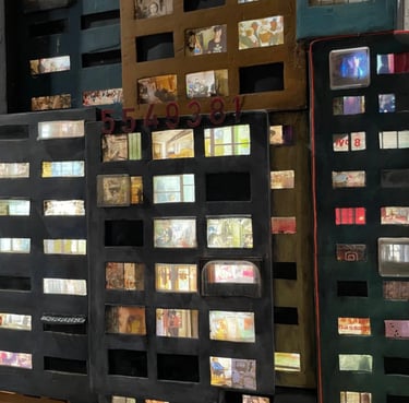 Mixed media art installation featuring miniature urban buildings with lit windows displaying photographs.