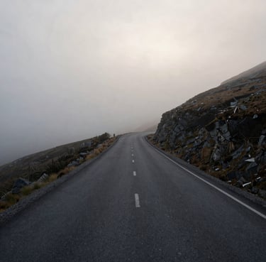 A minimalist wide-angle shot of a winding mountain road disappearing into a thick soft grey fog at dawn. The palette is muted with dark grey asphalt and off-white sky. Located in a rugged Global / Western landscape.