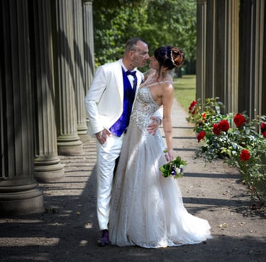 Ein Hochzeitspaar küsst sich unter einer Pergola mit Steinsäulen und einer Reihe roter Rosen; der Bräutigam trägt einen weiße