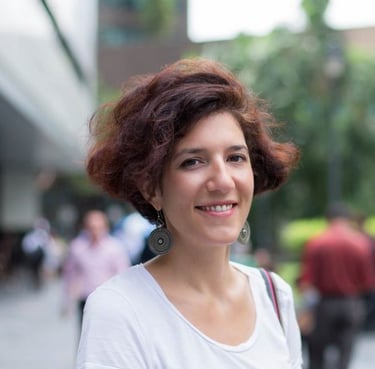 A smiling woman with short auburn curly hair wearing a white shirt and drop earrings outdoors.