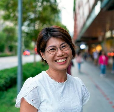 Smiling Asian woman with short hair and glasses wearing a white eyelet blouse on a city street.