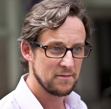 Professional man with brown hair and beard wearing tortoise shell glasses and a striped dress shirt.