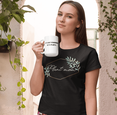 Woman holding a white plant-themed mug and wearing a black “Plant Mom” graphic t-shirt, standing near leafy green plants