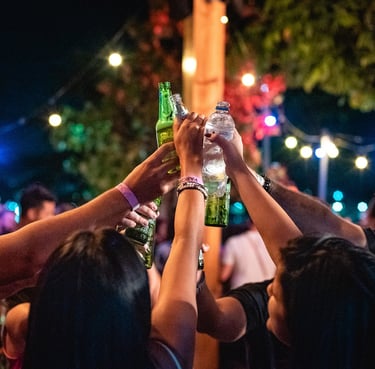 Friends toasting with beer bottles and water at an outdoor night festival with string lights.