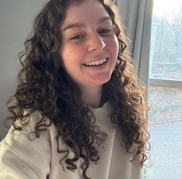 Smiling woman with long brown curly hair wearing a beige sweatshirt by a bright window.