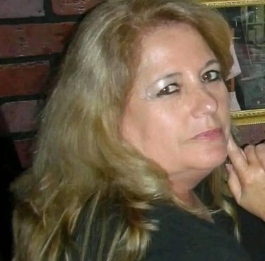 Close-up portrait of a woman with long blonde hair and makeup posing against a brick wall background.