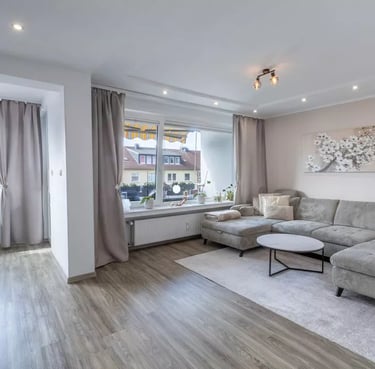 Modernes Wohnzimmer mit grauem Ecksofa, Holzboden und großen Fenstern mit neutralen Vorhängen.