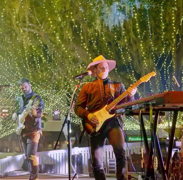 Musician performing live on stage under bright decorative string lights, playing electric guitar bes