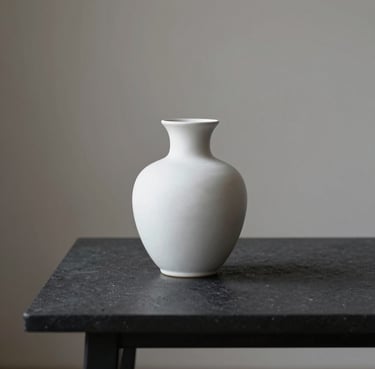 Minimalist still life of a single white ceramic vase on a dark charcoal table, soft side lighting, elegant Northern European home interior.