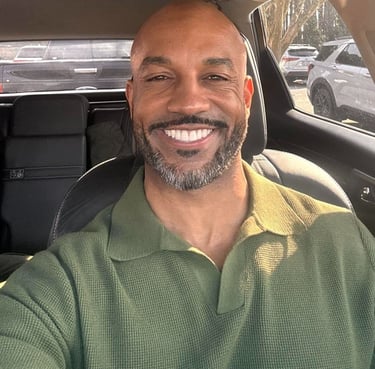 Smiling bald man with a salt and pepper beard wearing a green polo shirt inside a car.