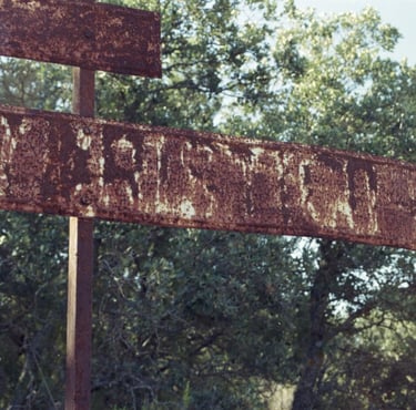 Cartel metálico oxidado con letras desgastadas frente a vegetación.