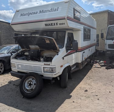  Old garage yard in rural Poland with campervan parked among broken-down cars and engine parts.