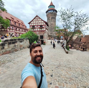 Smiling man taking a selfie at Nuremberg Castle with the Sinwell Tower and half-timbered houses.