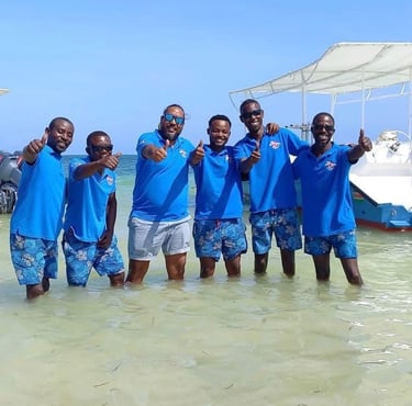 A group of smiling scuba intructors in blue shirts giving thumbs up in the ocean near boats.