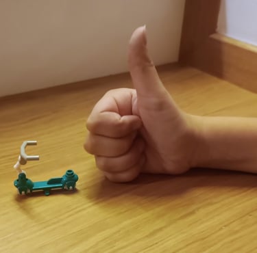 Dłoń dziecka z podniesionym do góry kciukiem, na biurku, obok maleńkiej hulajnogi z klocków.