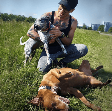 a man with tattoos playing with two happy dogs on the grass in a park