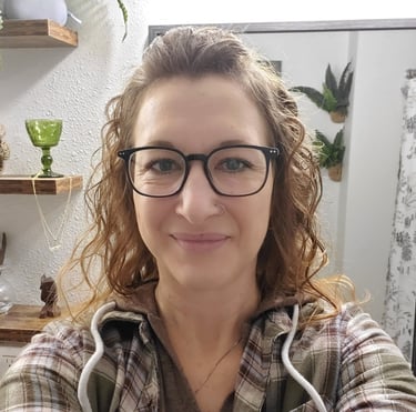 Woman with curly hair and black glasses wearing a plaid flannel shirt in a room with plants.