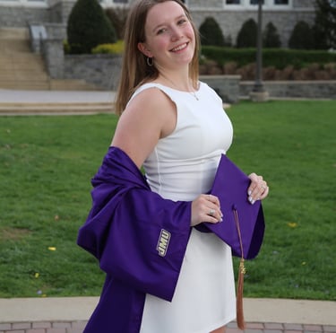 Smiling JMU graduate in a white dress holding a purple graduation cap and gown on campus.