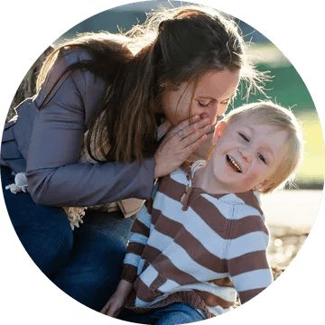 White woman with long, brown hair smiling and whispering into the ear of a blond-haired, smiling, white toddler boy