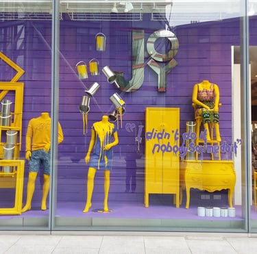 A vibrant purple and yellow retail window display featuring mannequins, paint buckets, and furniture.