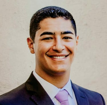 Foto do autor Leonardo Coelho sorrindo, com terno e parede branca ao fundo