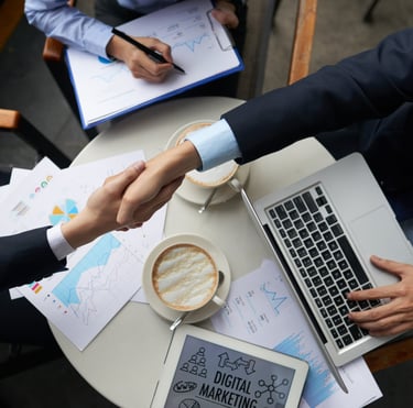 Business professionals shaking hands over a table with a laptop, digital marketing reports, and coffee.