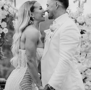 Smiling bride and groom in white wedding attire holding hands at an outdoor floral ceremony.
