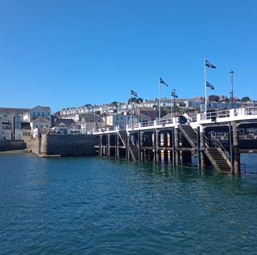 Concrete pier with flags in Falmouth, Cornwall, under a clear blue sky with coastal town houses.