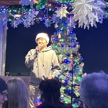 A man in a Santa hat performing holiday karaoke beside a lighted Christmas tree and festive decorations.