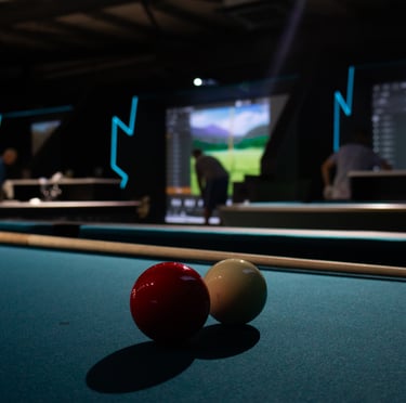 A pool table in the foreground with an indoor golf simulator bay behind it