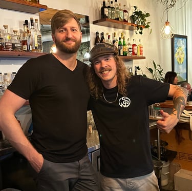 Two smiling male bartenders posing behind a well-stocked craft cocktail bar with liquor bottles on shelves.