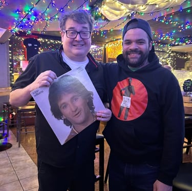 Two men smiling in a restaurant decorated with holiday lights while holding a Robin Williams vinyl record.