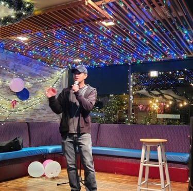 A male comedian performing stand-up comedy on a small stage with colorful festive lights and a microphone.