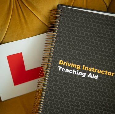 A spiral-bound driving instructor teaching aid manual resting next to a learner driver L plate on a yellow chair.
