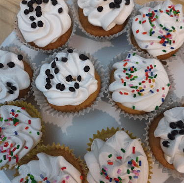 Freshly baked cupcakes with white swirl frosting topped with chocolate chips and colorful rainbow sprinkles.