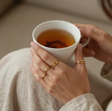 An elegant lifestyle photo of a woman's hands wearing delicate stacked gold-brass rings, holding a warm cup of herbal tea, with soft linen sleeves, cozy afternoon atmosphere in Brittany, France.